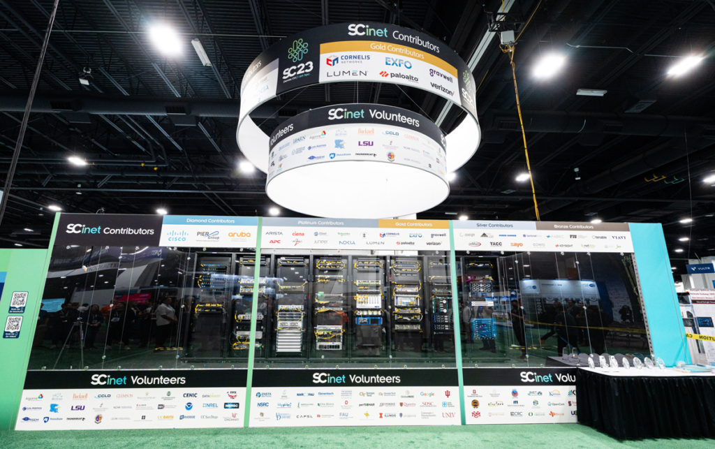 Hanging illuminated jumbotron displaying SCinet contributors and SCinet volunteers with racks of servers displayed underneath in an event booth.