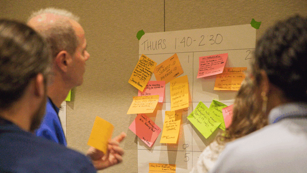 Four people stand in front of a large scheduling board labeled "THURS 1:40 - 2:30," reviewing a grid filled with colorful sticky notes.