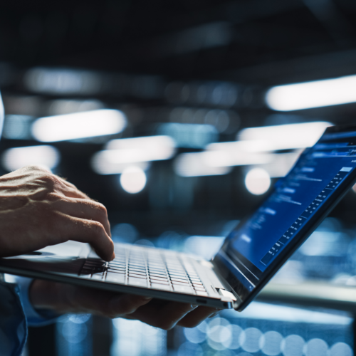 A close-up of a person in a light blue shirt holding and typing on a laptop in a high-tech server room or data center